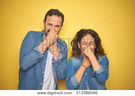 Beautiful middle age couple together wearing denim shirt over isolated yellow background smelling something stinky and disgusting, intolerable smell, holding breath with fingers on nose. Bad smells
