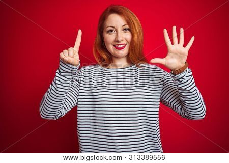 Young redhead woman wearing strapes navy shirt standing over red isolated background showing and pointing up with fingers number six while smiling confident and happy.