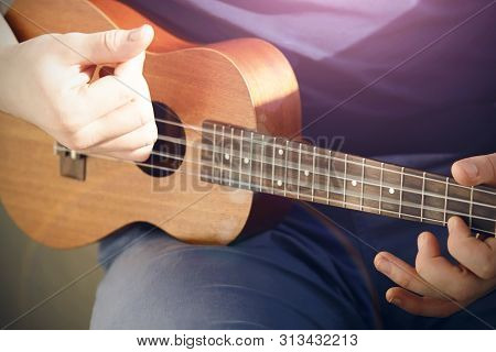 A Guy In A Blue T-shirt And Light Jeans Plays A Ukulele With Nylon Strings, Illuminated By Pleasant 