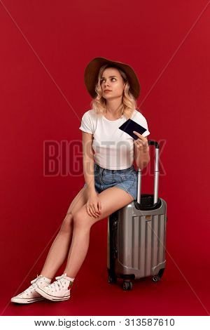 Exhaust Young Woman With Blod Curly Hair In A Sundown Hat Sits On A Grey Luggage Bag Holding Passpor