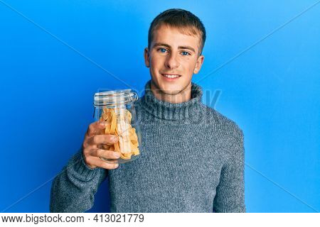 Young caucasian man holding jar of uncooked pasta looking positive and happy standing and smiling with a confident smile showing teeth 
