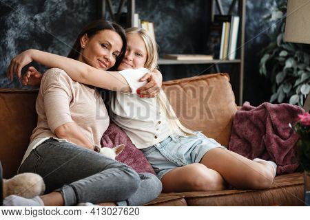 Mother and daughter relationship concept. A woman and a teenage girl are sitting in front of the TV on the couch and happy embarcing. The family spends time at home. Little dog lying on laps.