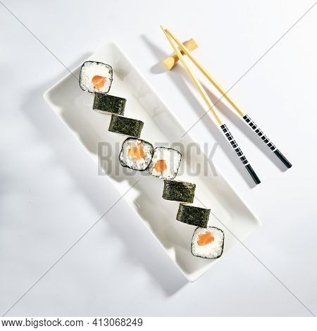Philadelphia Sushi Roll made with Smoked Salmon. Maki sushi with nori seaweed outside, salmon and cream cheese inside. White plate with wooden chopsticks on white table. Top view
