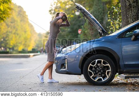 Puzzled Female Driver Standing On A Street Near Her Car With Popped Up Hood Inspecting Broken Engine