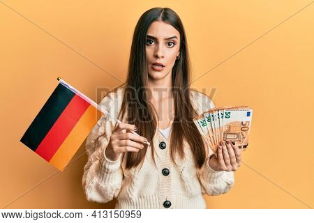 Beautiful brunette young woman holding germany flag and euros banknotes in shock face, looking skeptical and sarcastic, surprised with open mouth 