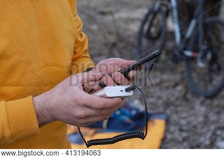 A Tourist Charges A Smartphone With A Power Bank On The Background Of A Backpack And A Bicycle In Na