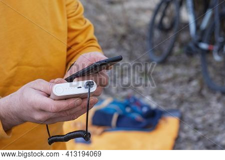 A Tourist Charges A Smartphone With A Power Bank On The Background Of A Backpack And A Bicycle In Na