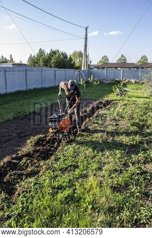 Male In The Spring Plows The Earth In Working Clothes With A Motoblock, Walk-behind Tractor