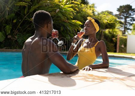 African american couple in water drinking drinks by poolside on sunny garden terrace. staying at home in isolation during quarantine lockdown.