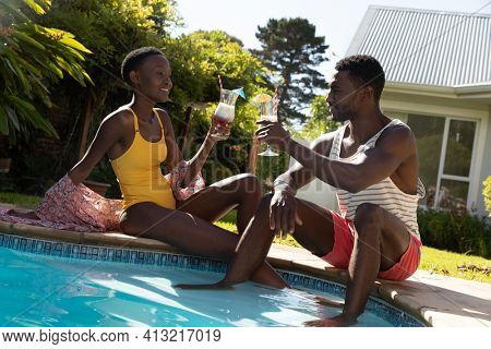 African american couple sitting at the poolside and drinking drinks on sunny garden terrace. staying at home in isolation during quarantine lockdown.