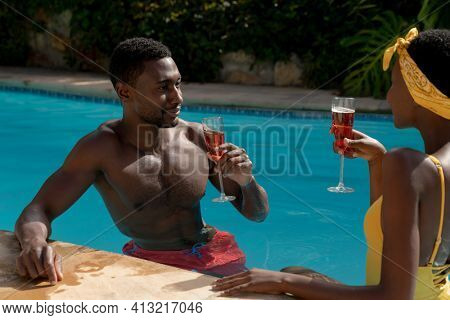 African american couple in water drinking drinks by poolside on sunny garden terrace. staying at home in isolation during quarantine lockdown.