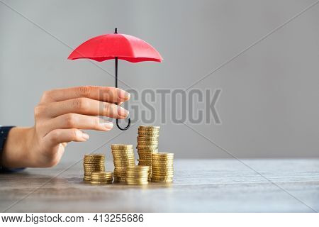 Young woman hand holding small red umbrella over pile of coins on table. Close up of stack of coins with female hands holding umbrella for protection. Financial safety and investment concept.  