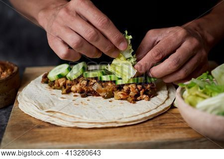 a young caucasian man fills a durum or a burrito, with chicken meat cooked with onion and red and green pepper, and fresh lettuce and some slices of raw cucumber