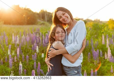 Young Mother Embracing Her Child Outdoor. Woman And Teenage Girl On Summer Field With Blooming Wild