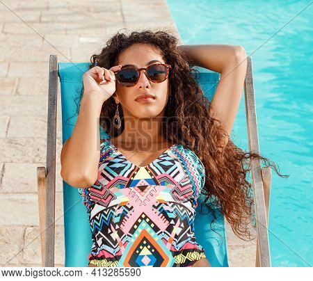 Young Caucasian Woman In Red Swimsuit With Tropical Cocktail Relaxing Near A Swimming Pool