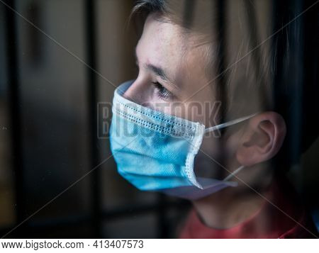Boy With A Medical Mask Looks Sadly Out Of The Window During His Confinement Because Of Convid 19