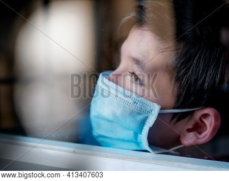 Boy With A Medical Mask Looks Sadly Out Of The Window During His Confinement Because Of Convid 19