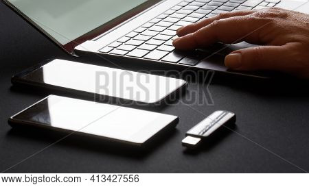 A Man Works At An Open Laptop Or Ultrabook Next To Two Smartphones And A Usb Flash Drive. Black Back