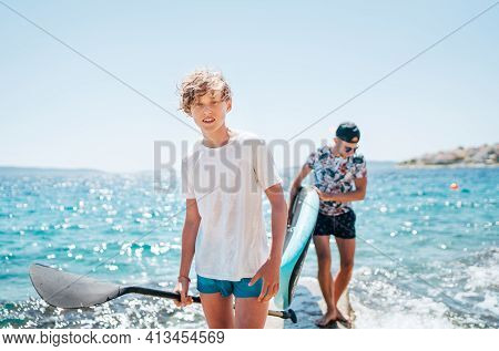 Smiling Blonde Teenage Son With A Paddle With Father Surfers Carrying The Stand-up Paddleboard On Th