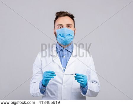 Young Doctor Wearing Medical Face Mask And Rubber Gloves, Holding Coronavirus Pcr Test Tube On Grey 