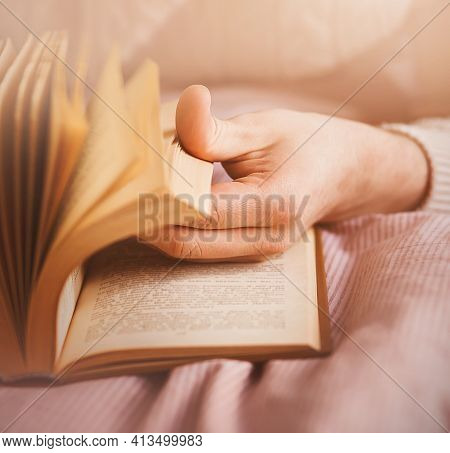 A Man In A White Sweater Flips Through An Interesting Old Book, Lying On A Bed With A Pink Striped B