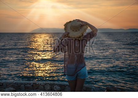 Woman In Straw Hat And Shorts By The Sea Looks At The Sunset