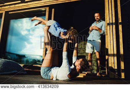 Family With Small Daughter Having Fun On Patio Of Wooden Cabin, Holiday In Nature Concept.