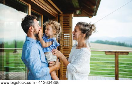 Family With Small Daughter On Patio Of Wooden Cabin, Holiday In Nature Concept.
