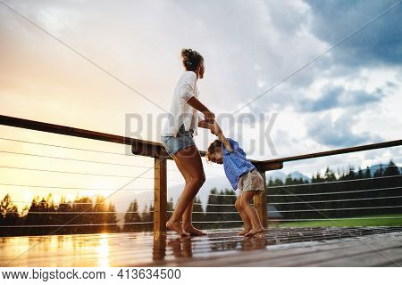 Mother With Small Daughter Playing In Rain On Patio Of Wooden Cabin, Holiday In Nature Concept.