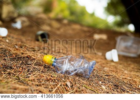 closeup of a used plastic bottle thrown on the ground of a forest, next to some other garbage, such as used food cans and plastic containers
