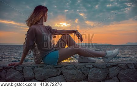 Woman Sits On Stones By The Sea And Looks At The Sunset