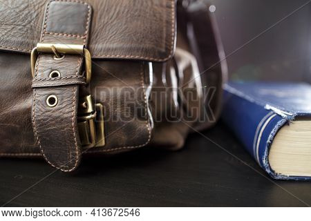 Old Leather Briefcase With Metal Buckles And A Closed Book Lying On The Table. Education Concept.