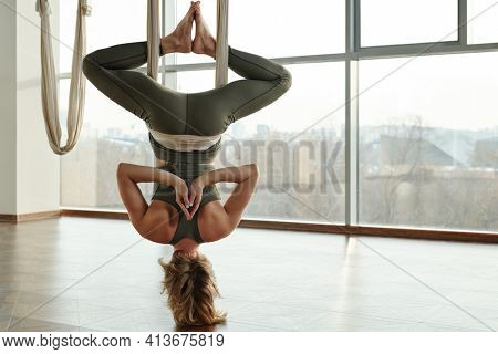 Back view of active and fit woman doing antigravity yoga exercise while hanging upside down with her hands and soles put together
