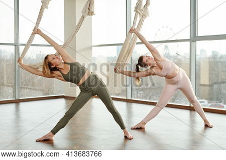 Two young active females in tracksuits using fly yoga equipment while doing sidebends on the floor of gym with their legs outstretched