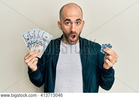 Young hispanic man playing poker holding dollars banknotes and chips afraid and shocked with surprise and amazed expression, fear and excited face. 