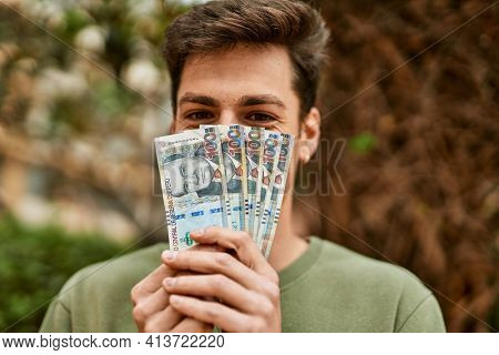 Young hispanic man smiling happy covering face with peruvian soles banknotes at the city.