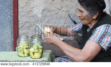 Wrinkled Senior Asian Woman Slices Raw Yellow Squash For Homemade Canning With Dill Seeds And Garlic