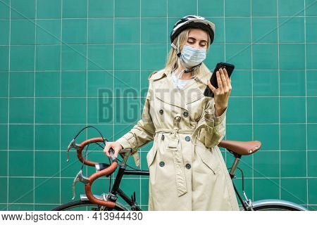 Woman in protective maskin, in helmet using her smartphone device. Female go to work on his sport retro bike. Girl standing on green background.