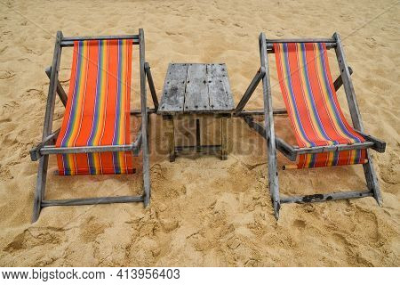 Two Empty Colorful Canvas Lounge Sling Chairs And Wooden Table On Sand Beach Of Resort In Thailand, 