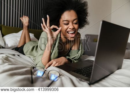 Happy Mid aged african woman using laptop computer while laying in bed at home, video cahtting while self isolating at home, ok gesture