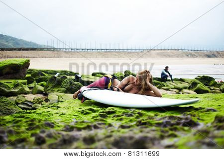 Professional surfer girl lying on moss rock against ocean horizon with her friend seat on background