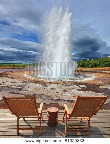  Geyser Strokkur in Iceland. Pillar of hot water and steam from forcing its way out of the ground. Two lounge chairs and  small table on  wooden platform for easy observation
