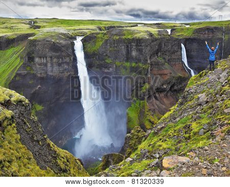 Spectacular waterfall Hayfoss in Iceland. Delighted woman - tourist in blue jacket standing on the edge of the cliff
