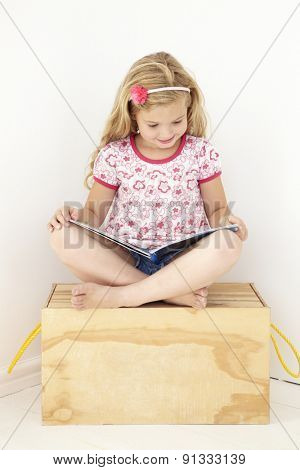 Young Girl Sitting On Wooden Toy Box Reading Book In Bedroom