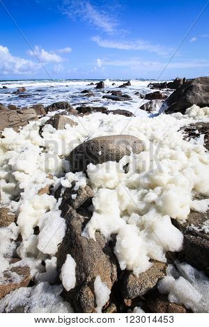Ocean foam congregating on the rocks as the tide goes out, against a big blue sky.