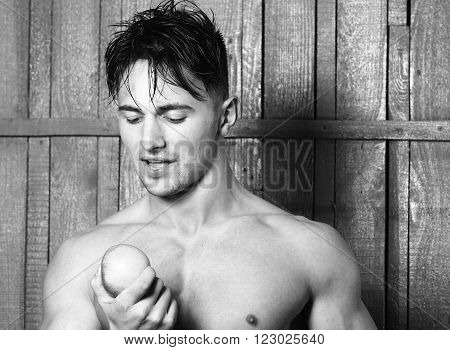 Sexy smiling emotional muscular young man with bare torso holding healthy foor of apple fruit standing indoor on wooden background, horizontal picture