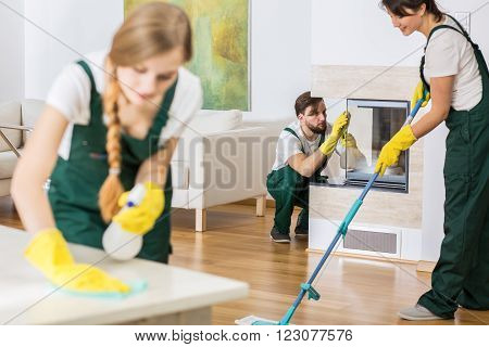 Three young professional cleaners in uniforms cleaning spacious living room