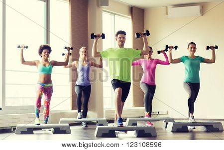 fitness, sport, aerobics and people concept - group of smiling people working out with dumbbells flexing muscles on step platforms in gym