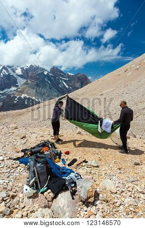 Group of Alpine Climbers Working on setting up Camping Tents with Many Gear Dropped Around and Rocky Mountain Moraine on Background