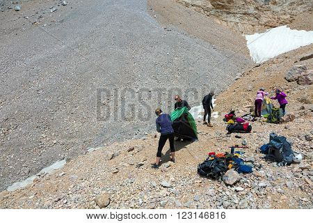 Group of Alpine Climbers Working on setting up Camping Tents with Many Gear Dropped Around and Rocky Mountain Moraine on Background